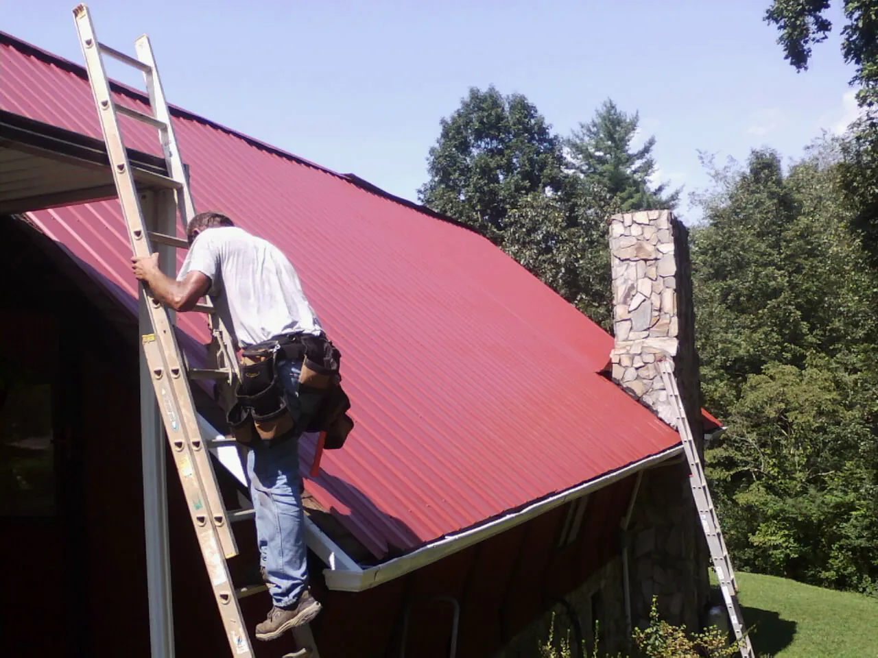 Roofer on a ladder beside a red metal roof and stone chimney — Soffit & Fascia Repair by Roofing Brothers in Athol